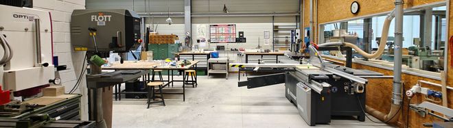 Großzügige Werkhalle mit stationären Holzbearbeitungsmaschinen, Arbeitstischen und Absauganlagen. Blick in einen technisch ausgestatteten Werkraum mit hohen Decken.