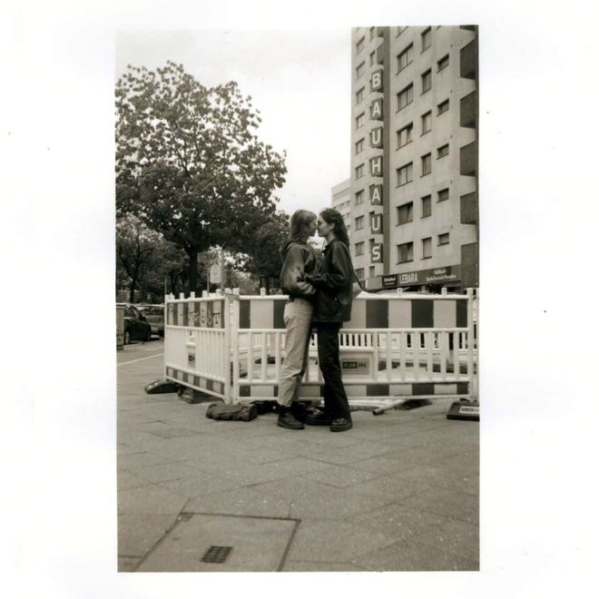 black-and-white photograph: Two women stand facing each other on a pavement as they begin to kiss. In the background are a residential building with a vertical "BAUHAUS" sign and temporary barriers.