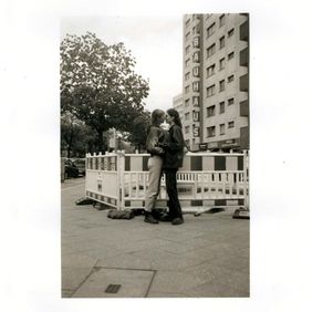»Herstory Repeats Itself«; Photo: Teresa Fischer black-and-white photograph: Two women stand facing each other on a pavement as they begin to kiss. In the background are a residential building with a vertical "BAUHAUS" sign and temporary barriers.