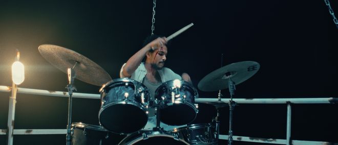 Film still from »DROWNING IS SILENT« by Alexander Knauer: an injured drummer plays on a platform reminiscent of a boat. The scene is lit in a dark, cinematic atmosphere.