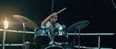 Film still from »DROWNING IS SILENT« by Alexander Knauer: an injured drummer plays on a platform reminiscent of a boat. The scene is lit in a dark, cinematic atmosphere.