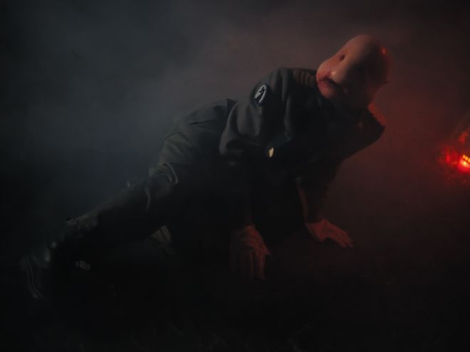 Person in Uniform mit glatter, puppenartiger Gesichtsform bewegt sich am Boden in dunkler, nebliger Umgebung mit rotem Licht im Hintergrund.