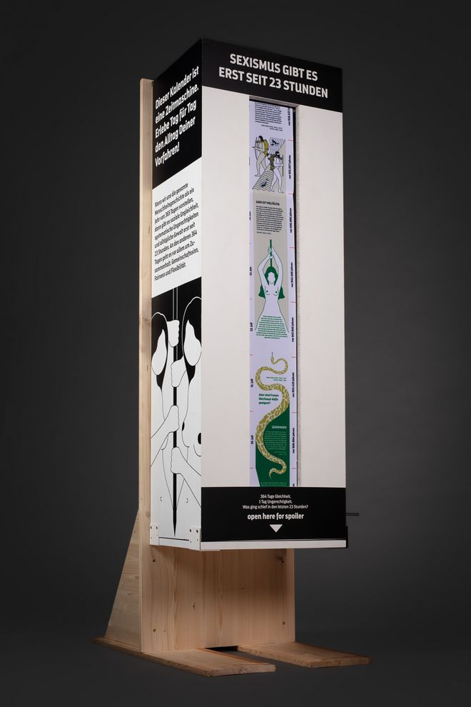 Isolated view of the mechanical crank station against a dark background. A narrow, colourfully illustrated fabric strip runs through a viewing window in the tall wooden structure; a short introductory text is displayed on the side.