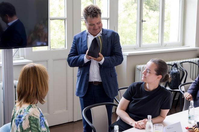 Jürgen Dusel steht im Besprechungsraum und hält mit Freude ein Geschenk in der Hand, das ihm überreicht wurde. Zu sehen sind seine Kolleginnen, die ihn anschauen und am Tisch sitzen.