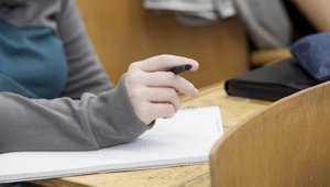 Bild im Hörsaal mit Notizblock und Hand mit Stift im Fokus
