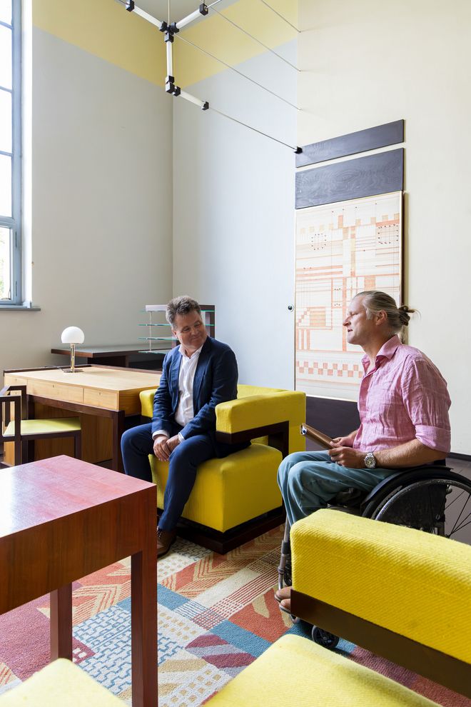 Jürgen Dusel und Martin Ahner im Gropiuszimmer. Dusel sitzt auf einem von Gropius' Sesseln, Ahner neben ihm und erzählt etwas. Beide sehen sehr positiv gestimmt aus, Dusel lächelt. Um sie herum ist das Gropiuszimmer zu erkennen: der Schreibtisch mit der Wagenfeldlampe, die Sessel, ein kleines Tischchen, hinten ein Wandteppich.