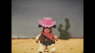 Knetfigur eines Schweins mit pinkem Cowboyhut steht in einer sandigen Wüstenlandschaft mit Kakteen im Hintergrund.