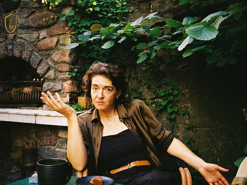 Portrait of Polina Horošina sitting at a table in a leafy courtyard, looking at the camera with her hands outstretched.