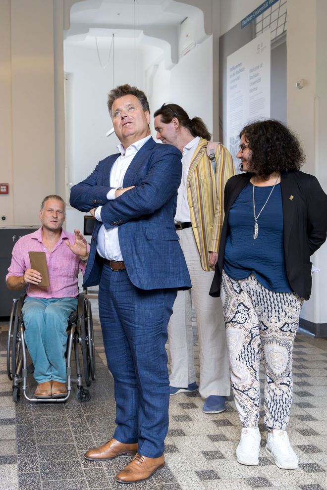 Jürgen Dusel steht mit zwei Mitarbeitern und einer Mitarbeiterin im Foyer des Hauptgebäudes. Martin Ahner berichtet über etwas, das sie sich anschauen, auf dem Foto jedoch nicht zu sehen ist. Die anderen drei hören ihm gespannt zu. Dusels Blick geht raus aus dem Bild, wahrscheinlich zu dem Objekt, das beschrieben wird. Er steht im Zentrum des Fotos.