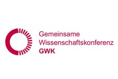 The type-logo shows in red print on a white background the words: »Gemeinsame Wissenschaftskonferenz GWK« (right-hand side). On the left-hand side of the logo, there's a circle drawn in red lines. The left half of the circle consists of a dotted line, the right half of a solid line.