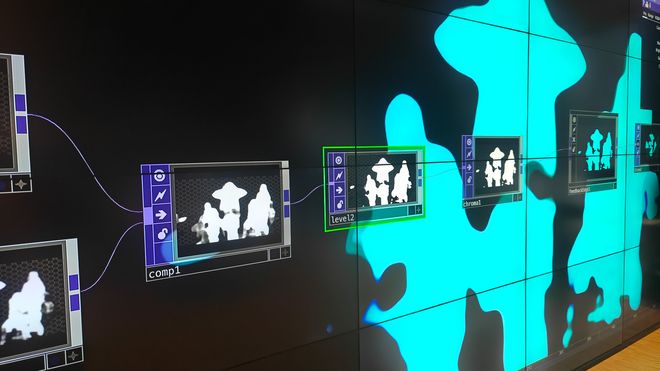 Mehrere miteinander verbundene Module der Software TouchDesigner sind auf einer LED-Wand dargestellt. Daneben erscheinen abstrahierte Silhouetten von Personen.