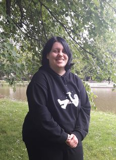 Jasmin Rogge hat kinnlange schwarze Haare und dunkle Augen. Sie trägt einen schwarzen Kapuzenpulli und eine schwarze Hose. Auf dem Pulli ist das Bild eines liegenden Astronauten zu sehen, der einen Papierflieger in die Luft wirft. Sie lächelt freundlich in die Kamera.