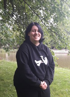 Jasmin Rogge has chin-long black hair and dark eyes. She is wearing a black hoodie and black pants. On the hoodie, there's the graphic of an astronaut, who is lying on his side while throwing a paper plane into the air. She is smiling at the camera.