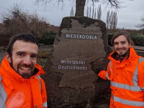 Zwei Männer stehen vor einem großen Gedenkstein, zeigen auf ihn und blicken direkt in die Kamera.
