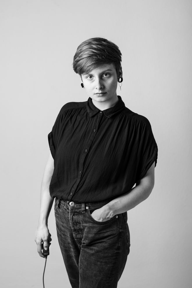 Das Schwarz-Weiß-Foto zeigt Konzeptkünstlerin Larissa Barth. Barth hat kurze, dunkelblonde Haare und schwarze Tunnel-Ohrringe. Sie trägt eine dunkle Jeans und eine Bluse aus dickem, schwarzem Stoff mit kurzen Ärmeln. Ihre linke Hand steckt in ihrer Hosentasche. Die rechte Hand hält den Selbstauslöser. Sie blickt selbstbewusst in die Kamera.