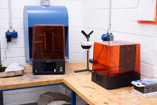 Zwei Harz-3D-Drucker mit orangefarbenen Schutzhauben stehen nebeneinander auf einer Werkbank in der Kunststoffwerkstatt.