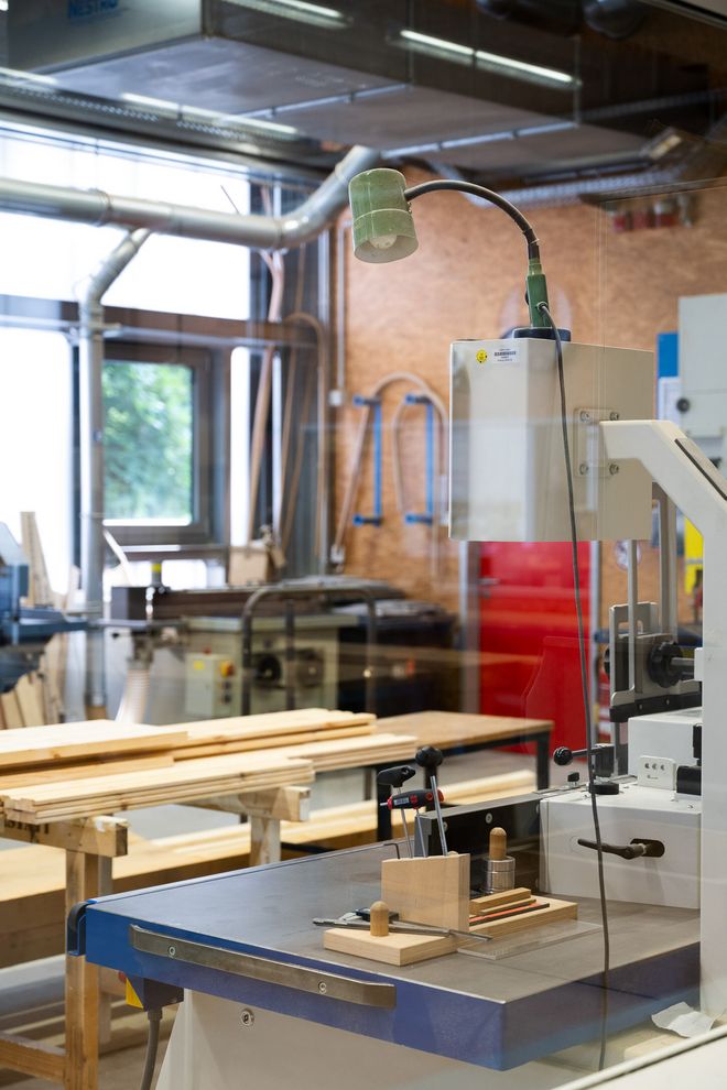 Detail view of a stationary machine in a woodworking workshop with workpieces and hand tools on the work surface. Additional machines are visible in the background.