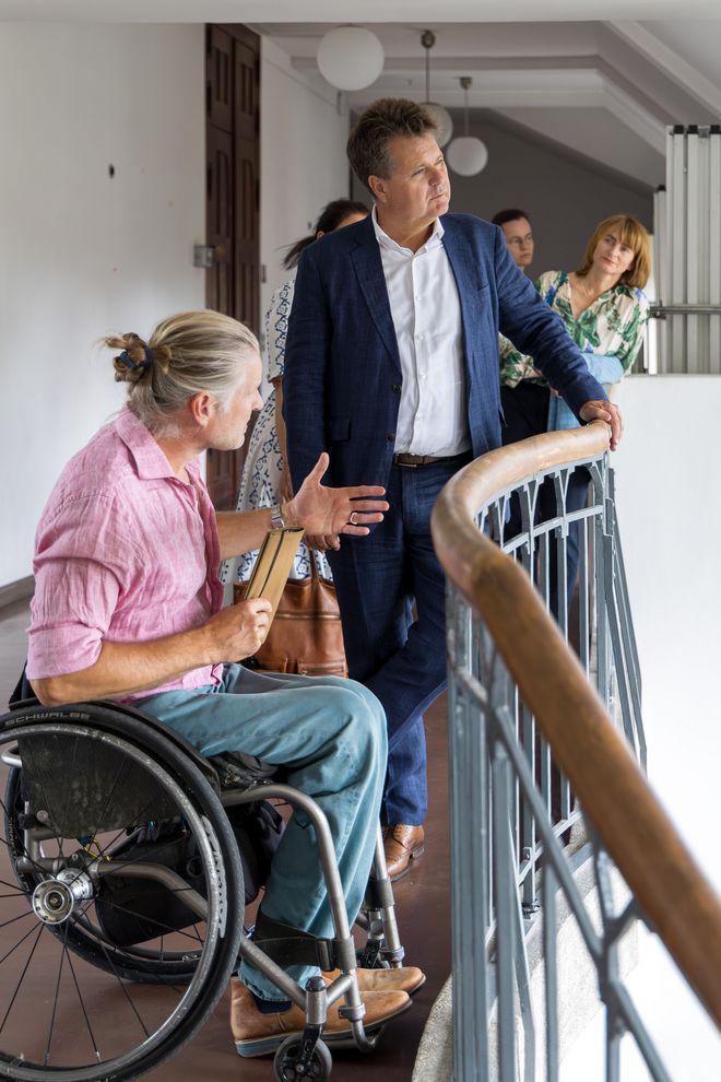Jürgen Dusel mit Martin Ahner im obersten Stock im Hauptgebäude, sie sind im Flur. Dusel hat eine Hand am Treppengeländer, Ahner sitzt neben ihm im Rollstuhl und erzählt etwas, dabei gestikuliert er mit seinen Händen. Im Hintergrund hören weitere Mitarbeiterinnen von Dusel und der Universität zu.