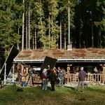 Filmset von »Raíces« vor einer Holzhütte im Wald. Das Filmteam steht mit Kamera-, Licht- und Tontechnik vor einer Hütte.