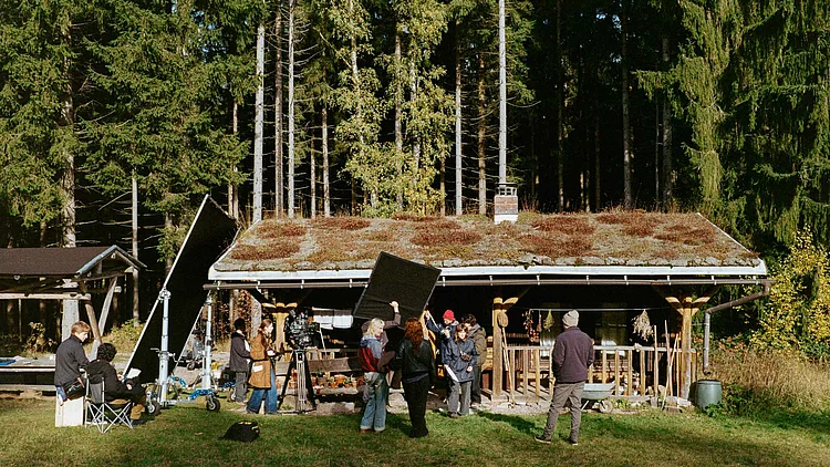 The film set for *Raíces* in front of a wooden hut in the woods. The film crew is standing in front of the hut with their camera, lighting and sound equipment.