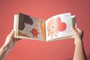 Book view of »RED AF* – Red Asian Flush« by Soo Ah Youn and Anastasia Fast: two hands hold an open illustrated book against a red background. The pages show the character »Blush« and a message about listening to one’s body.