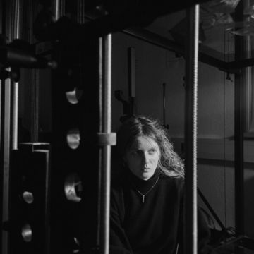 Schwarz-Weiß-Fotografie von Anya Ryzhkova, die zwischen vertikalen Metallstangen und technischen Bauteilen sitzt. Die Person blickt konzentriert zur Seite; die Umgebung wirkt wie ein Labor oder eine technische Installation.