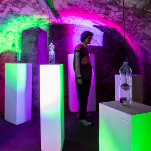 Eine Person bewegt sich durch einen gewölbten, steinernen Ausstellungsraum mit grüner und magentafarbener Lichtinszenierung. Auf leuchtenden weißen Sockeln stehen transparente PET-Flaschen, in denen kleine Lautsprecher hängen; Kabel und technische Elemente sind sichtbar. Die Szene zeigt eine interaktive Installation.