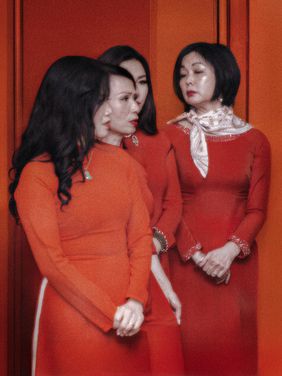 »Every Weekend, Our Attic Becomes Vietnam«; Photo: Mai Do Three women wearing red áo dài stand in front of an orange wall. The woman on the right rests her hand on another woman's shoulder while all three look in different directions.