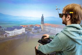 Das Bild zeigt einen Studenten, der eine VR-Brille trägt und auf eine dreidimensionale Visualisierung von Weimar schaut. Er navigiert sich durch die Landschaft mittels eines kugelförmigen Eingabegerätes.