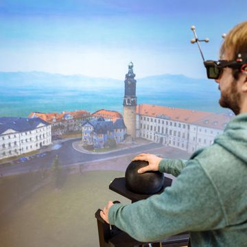 Das Bild zeigt einen Studenten, der eine VR-Brille trägt und auf eine dreidimensionale Visualisierung von Weimar schaut. Er navigiert sich durch die Landschaft mittels eines kugelförmigen Eingabegerätes.