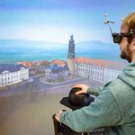 Das Bild zeigt einen Studenten, der eine VR-Brille trägt und auf eine dreidimensionale Visualisierung von Weimar schaut. Er navigiert sich durch die Landschaft mittels eines kugelförmigen Eingabegerätes.