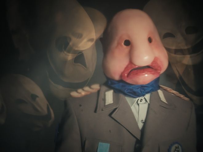 Person in Uniform mit glatter, puppenartiger Gesichtsform steht vor mehreren hellen Gesichtern im Hintergrund.