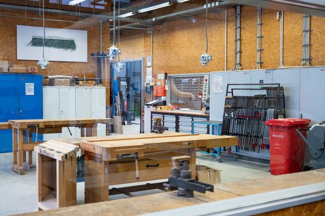 View through a glass pane into a woodworking shop with workbenches, cabinets and stationary machines. Tools are neatly arranged on the wall.