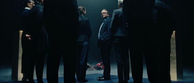 Film still from »DROWNING IS SILENT« by Alexander Knauer: a group of people in suits stands in a dark room. In the background, a person in red and white clothing lies on the floor.