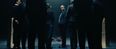 Film still from »DROWNING IS SILENT« by Alexander Knauer: a group of people in suits stands in a dark room. In the background, a person in red and white clothing lies on the floor.