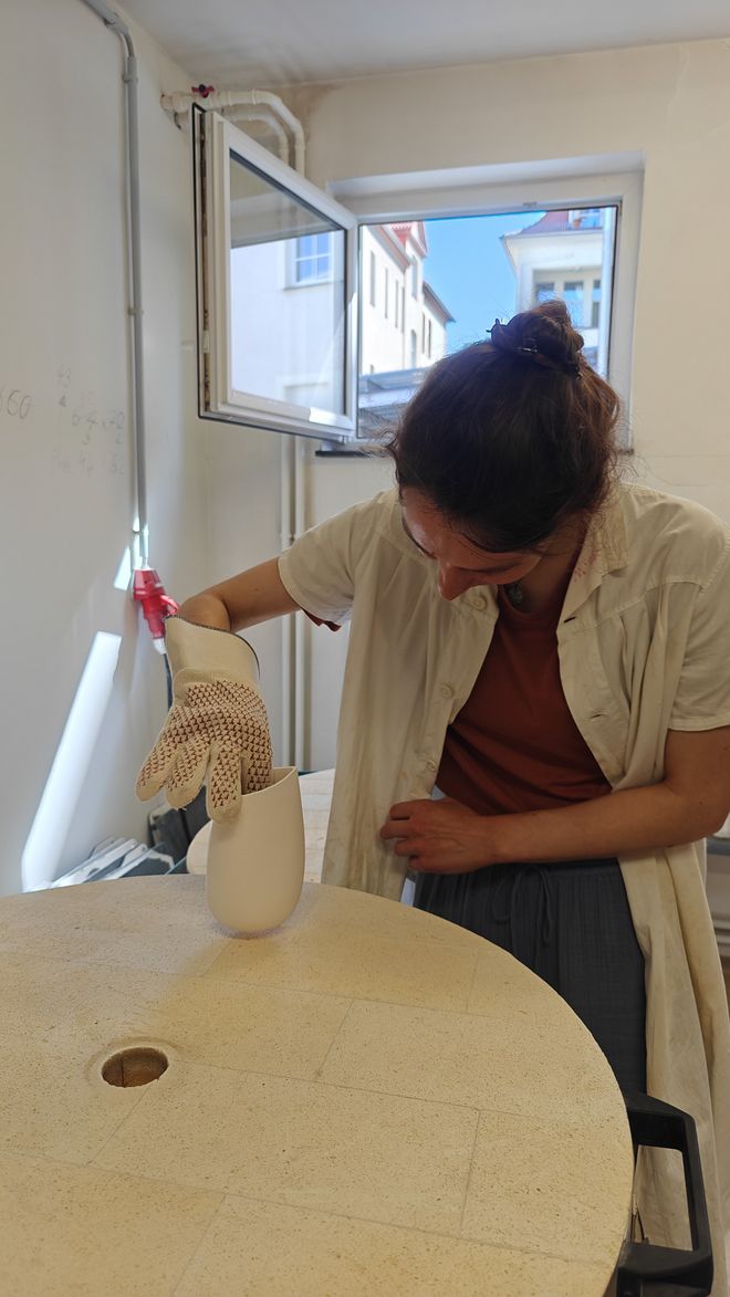 Charlotte setzt mit einem hitzebeständigen Handschuh ein keramisches Gefäß vorsichtig in einen Brennofen.