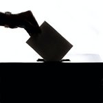 Eine Hand wirft einen Wahlzettel in eine Wahlurne ein.