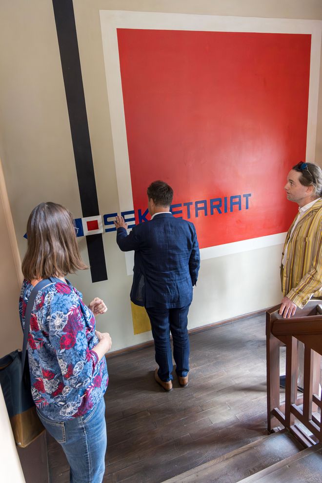 Christiane Hempel, Jürgen Dusel und Gunter Seidel stehen in einem schmaleren Treppenhaus des Hauptgebäudes vor einer großflächigen Wandmalerei. Hempel und Seidel mit etwas Abstand, Dusel steht direkt vor der Wand und betastet die Kunstmalerei. Es handelt sich um die Wandmalerei Quadrat von Herbert Bayer, rekonstruiert 1976 von Werner Claus, eine Mischtechnik. Es is eine große quaddratische rote Fläche, mit einem breiten weißen Rand umrundet. Links daneben mit etwas Abstand ein vertikaler schwarzer langer, breiter Balken. Ein dunkelblauer Schriftzug Sekretariat ragt links anfangend vom Balken hinein in das rote Quadrat. Links davon ist ein blauer Pfeil, der den Weg weit. Bayer (1900 – 1985) studierte von 1921 bis 1925 am Staatlichen Bauhaus in Weimar. Er entwarf die Schrift für den Einband des »Bauhaus-Manifest«. Die Wandmalereien im Nebentreppenhaus entstanden anlässlich der großen Bauhaus-Ausstellung 1923 und zeigen die geometrischen Grundformen Kreis, Quadrat sowie Dreieck, die Walter Gropius als verbindendes Thema für die Gestaltung im Hauptgebäude vorgegeben hatte. 