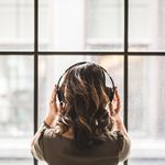 Eine Frau vin hinten steht vor einem großen Fenster, ihre Hände liegen auf ihrem Kopfhöhrer, den sie auf hat