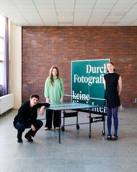 Tarek Rishmawi, Luisa Hörning und Marleen Kölmel stehen und sitzen an einer Tischtennisplatte vor dem Ausstellungsmotiv »Durch Fotografie keine Einsichten«