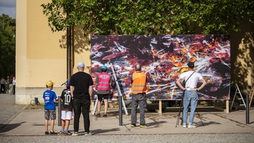 einige Menschen beobachten das Ankelben eines neuen Großplakates an einer Aufstellwand neben dem Hauptgebäude anlässlich der summaery