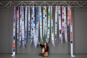 A 55-metre-long printed fabric panel from the work »Sexismus gibt es erst seit 23 Stunden«; photo: Anne-Kathrin Lutherberg and Annika Daub Exhibition view of »Sexismus gibt es erst seit 23 Stunden.« by Nina Buschendorf: a 55-metre-long, colourfully printed fabric strip hangs in multiple loops from a metal structure. Nina Buschendorf is sitting beneath it and touching one of the strips.