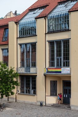 Das Foto zeigt ein Banner mit der »Inter*-inklusiven Progress-Pride-Flagge« am zentralen Balkon des Hauptgebäudes der Bauhaus-Universität Weimar.