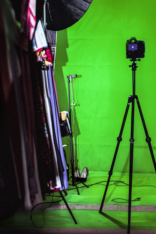 A camera on a tripod stands in front of a green screen. Stage costumes hang in the foreground, with studio lighting and additional equipment visible nearby.