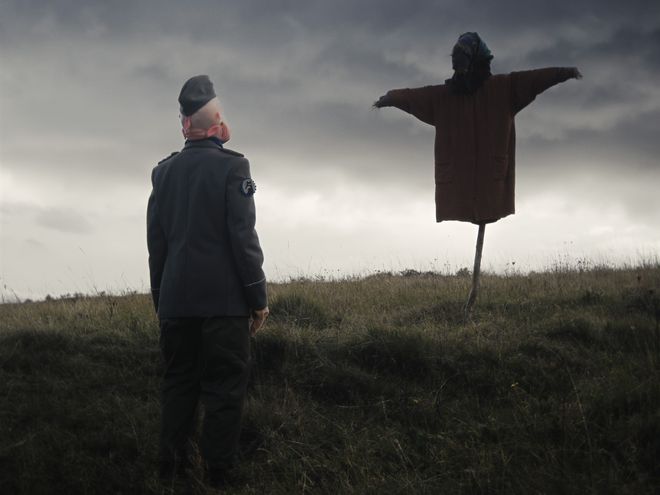 Person in Uniform steht auf einem Feld und blickt zu einer Vogelscheuche auf einem Hügel.