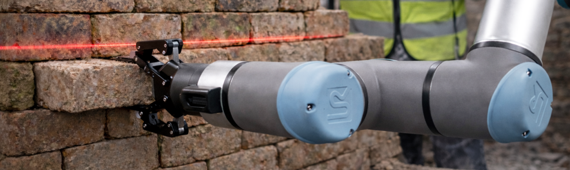 Roboterarm mit Greifwerkzeug arbeitet an einer Mauer aus Ziegelsteinen. Ein roter Laserstrahl markiert die Ausrichtung, im Hintergrund ist eine Person mit Warnweste unscharf zu erkennen.
